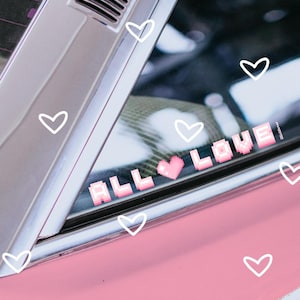 Puede incluir: Un coche rosa con una pegatina blanca que dice "ALL LOVE" en letras pixeladas con un corazón rosa en el medio. La pegatina está en la ventana trasera del coche. Hay corazones blancos esparcidos alrededor de la pegatina.