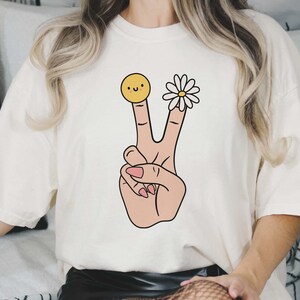 May include: White t-shirt with a peace sign graphic. The peace sign is made up of two fingers with a yellow smiley face on one finger and a white daisy on the other.