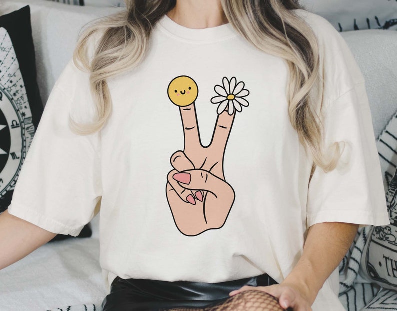 May include: White t-shirt with a peace sign graphic. The peace sign is made up of two fingers with a yellow smiley face on one finger and a white daisy on the other.