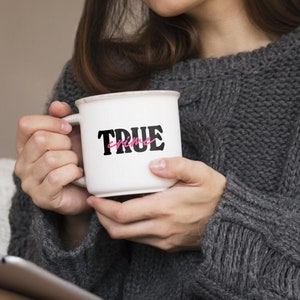 May include: White ceramic mug with black text that reads "TRUE crime" with the word "crime" in pink. The mug is being held by a person wearing a gray sweater.