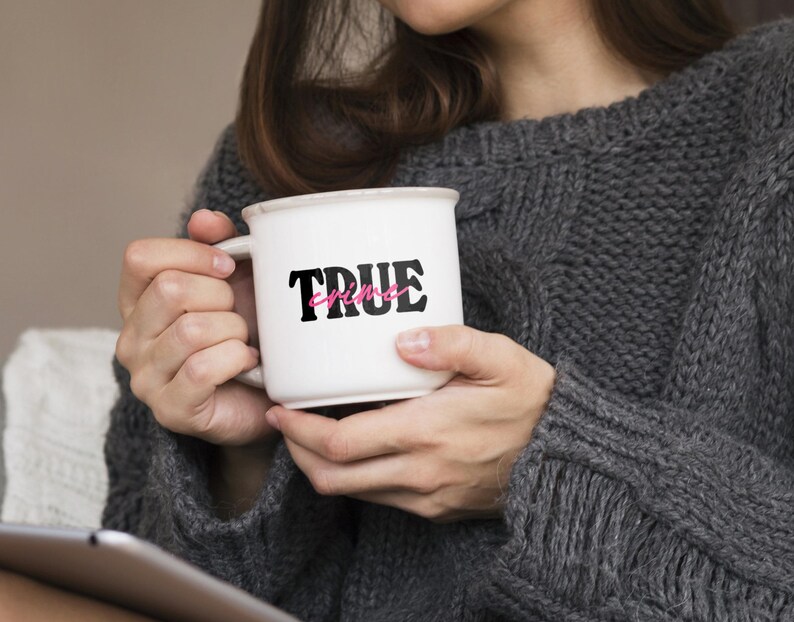 May include: White ceramic mug with black text that reads "TRUE crime" with the word "crime" in pink. The mug is being held by a person wearing a gray sweater.