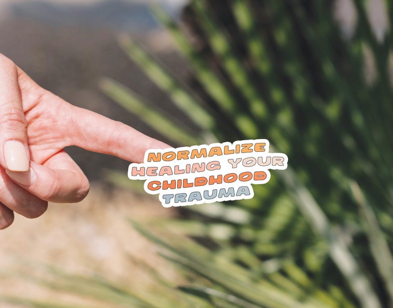 May include: A sticker with the text "Normalize Healing Your Childhood Trauma" in a rainbow color scheme. The sticker is on a blurred background of green foliage.