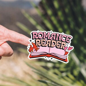 May include: A pink and red sticker with the text "Romance Reader" and a stylized image of a book with flames. The sticker has two small hearts on the right side.