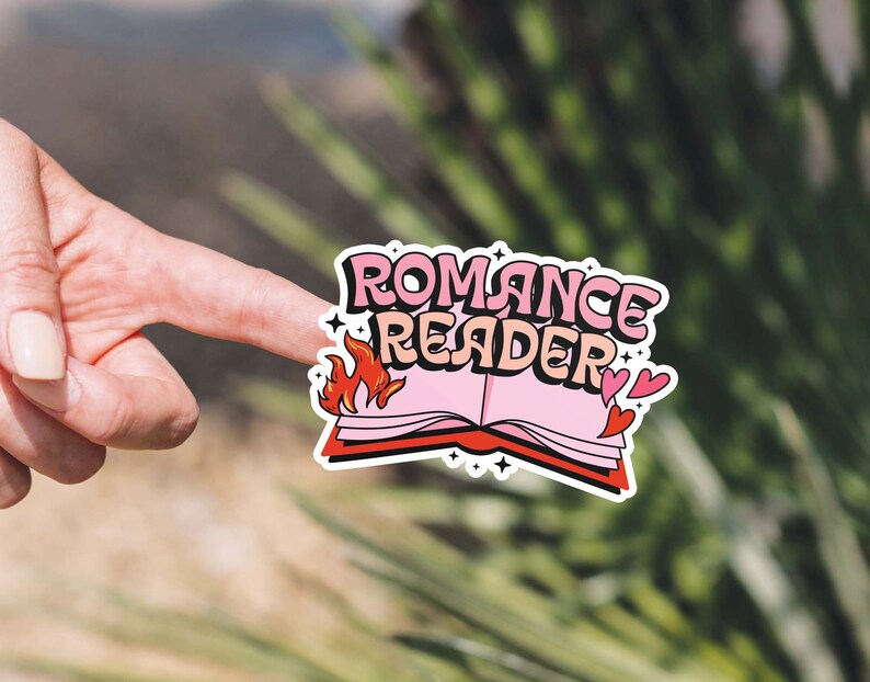 May include: A pink and red sticker with the text "Romance Reader" and a stylized image of a book with flames. The sticker has two small hearts on the right side.