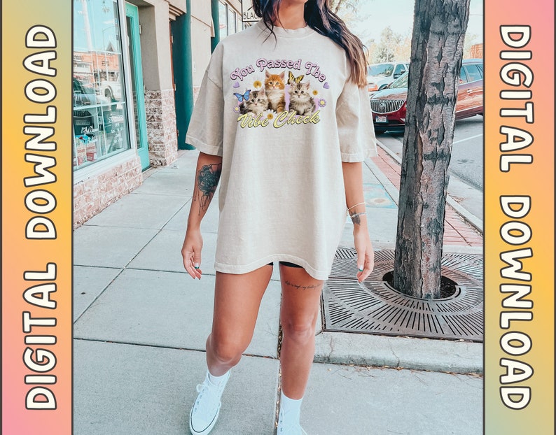 Op de afbeelding: Een beige oversized t-shirt met een afbeelding van kittens en de tekst "You Passed The Vibe Check".