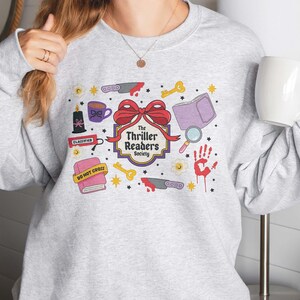 May include: A light gray sweatshirt featuring a colorful graphic design. The design includes a red bow, knives, keys, a book, a magnifying glass, a handprint, and the text "The Thriller Readers Society".