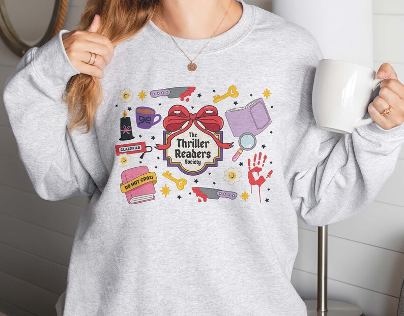 May include: A light gray sweatshirt featuring a colorful graphic design. The design includes a red bow, knives, keys, a book, a magnifying glass, a handprint, and the text "The Thriller Readers Society".