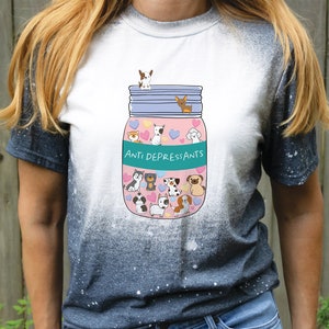 Pu&ograve; includere: Una maglietta bianca con effetto tie-dye, che presenta un'illustrazione di cartoni animati di un barattolo di vetro pieno di cani di cartoni animati e cuori. Il barattolo ha un'etichetta turchese che dice "Anti Depressants".