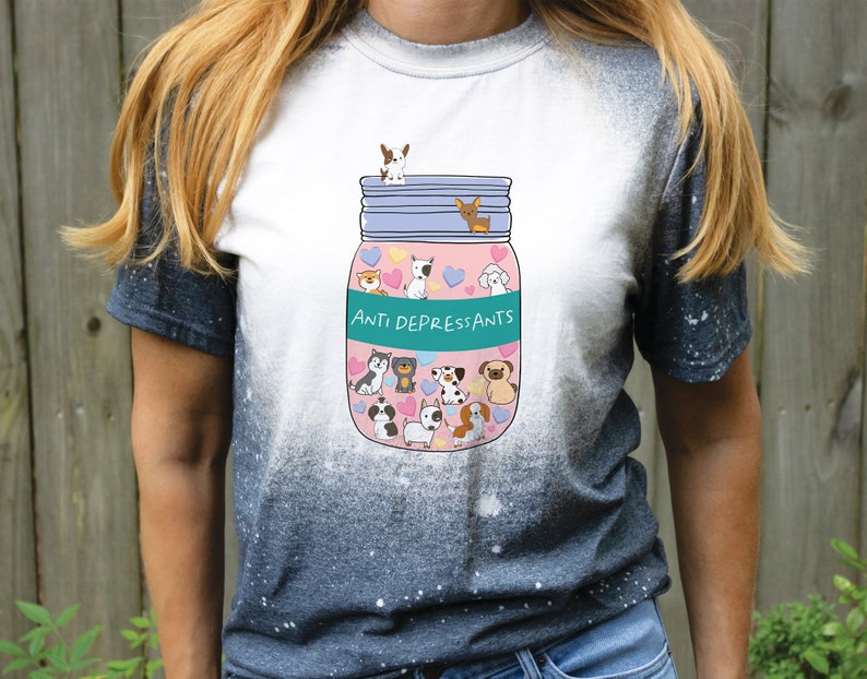 Pu&ograve; includere: Una maglietta bianca con effetto tie-dye, che presenta un'illustrazione di cartoni animati di un barattolo di vetro pieno di cani di cartoni animati e cuori. Il barattolo ha un'etichetta turchese che dice "Anti Depressants".