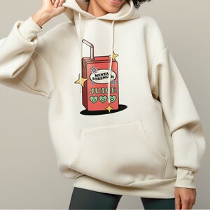 Puede incluir: Una sudadera con capucha de color crema con un gr&aacute;fico de una caja de jugo roja con las palabras "Mental Strength Juice" y tres corazones verdes. La caja de jugo tiene una pajita que sobresale y est&aacute; rodeada de estrellas amarillas.