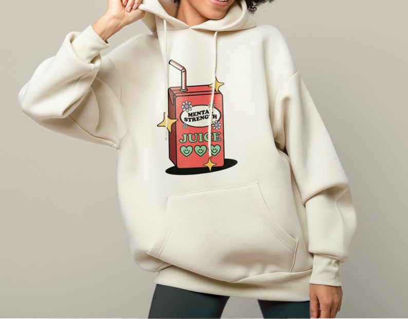 Puede incluir: Una sudadera con capucha de color crema con un gr&aacute;fico de una caja de jugo roja con las palabras "Mental Strength Juice" y tres corazones verdes. La caja de jugo tiene una pajita que sobresale y est&aacute; rodeada de estrellas amarillas.