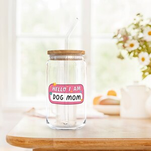 Peut inclure: Un bocal en verre transparent avec un couvercle en bois et une paille. Le bocal a un autocollant rose qui dit "Hello I am Dog Mom".