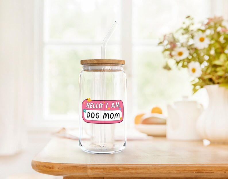 Peut inclure: Un bocal en verre transparent avec un couvercle en bois et une paille. Le bocal a un autocollant rose qui dit "Hello I am Dog Mom".
