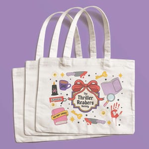 May include: Three white tote bags with cream-colored handles. Each bag features a colorful design with a bow, knives, keys, a magnifying glass, a handprint, and the text "The Thriller Readers Society."