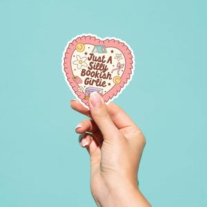 May include: A heart-shaped sticker with a pink scalloped border and the text "Just A Silly Bookish Girlie." The sticker features illustrations of a tent, flowers, a strawberry, and a book. Held by a hand against a teal background.