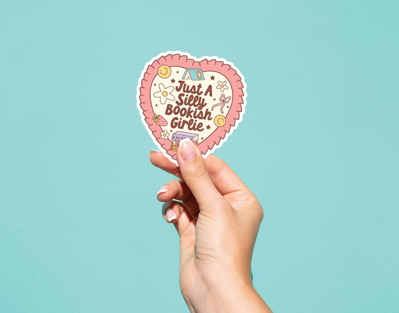 May include: A heart-shaped sticker with a pink scalloped border and the text "Just A Silly Bookish Girlie." The sticker features illustrations of a tent, flowers, a strawberry, and a book. Held by a hand against a teal background.