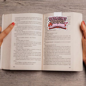 May include: A close-up of an open book with a pink and white bookmark that says "Romance Reader". The bookmark features a stylized open book with hearts and flames. The book is open to a page with Spanish text.