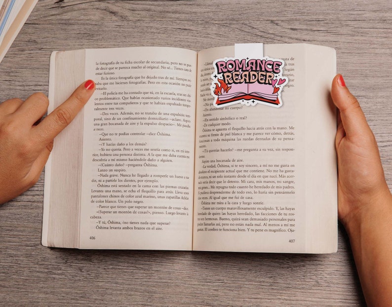 May include: A close-up of an open book with a pink and white bookmark that says "Romance Reader". The bookmark features a stylized open book with hearts and flames. The book is open to a page with Spanish text.