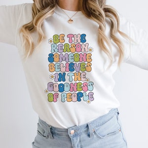 May include: White sweatshirt with the text "Be the reason someone believes in the goodness of people" in colorful, bubble-like letters.