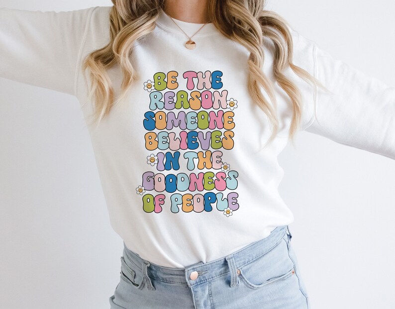 May include: White sweatshirt with the text "Be the reason someone believes in the goodness of people" in colorful, bubble-like letters.