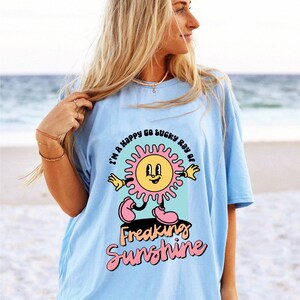 Op de afbeelding: Een lichtblauw t-shirt met een afbeelding van een lachende zon met roze stralen en de tekst "I'm a happy go lucky ray of freaking sunshine".