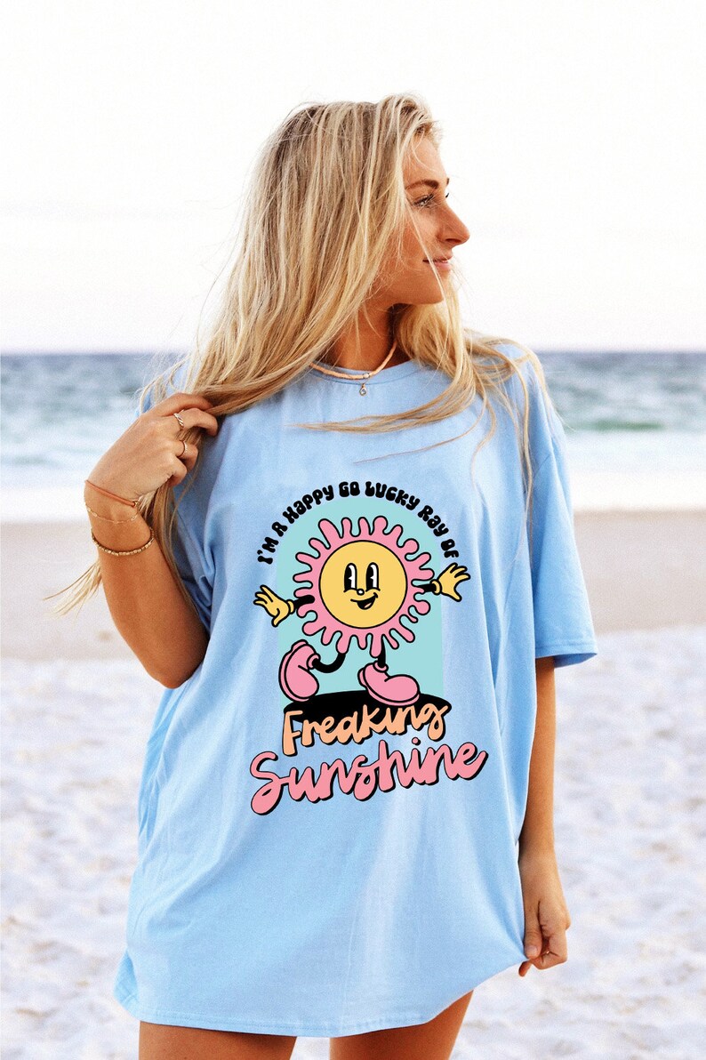 Op de afbeelding: Een lichtblauw t-shirt met een afbeelding van een lachende zon met roze stralen en de tekst "I'm a happy go lucky ray of freaking sunshine".