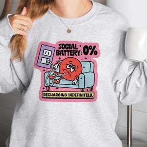May include: A light gray sweatshirt featuring a graphic of a red character with a sad face, lounging on a blue couch. The graphic includes the text "SOCIAL BATTERY: 0%" and "RECHARGING INDEFINITELY."