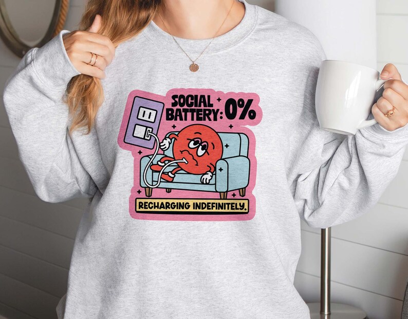 May include: A light gray sweatshirt featuring a graphic of a red character with a sad face, lounging on a blue couch. The graphic includes the text "SOCIAL BATTERY: 0%" and "RECHARGING INDEFINITELY."