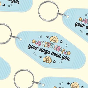 May include: Light blue key chain with a white and pink design. The keychain features a cartoon dog and the text "Drive Safe, your dogs need you".