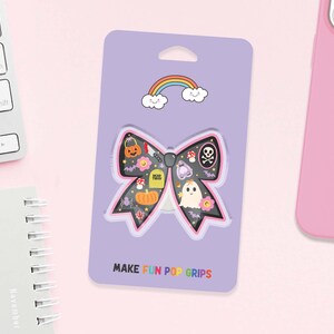 May include: A purple card with a bow-shaped phone grip featuring Halloween illustrations. The bow has a black background with a skull, pumpkin, ghost, and other spooky elements. The text "MAKE FUN POP GRIPS" is at the bottom, and a rainbow is at the top.