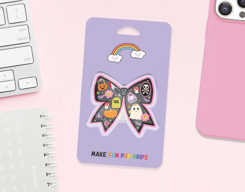 May include: A purple card with a bow-shaped phone grip featuring Halloween illustrations. The bow has a black background with a skull, pumpkin, ghost, and other spooky elements. The text "MAKE FUN POP GRIPS" is at the bottom, and a rainbow is at the top.