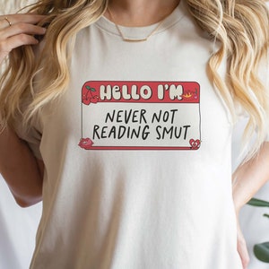 May include: White t-shirt with a red name tag that says "Hello I'm Never Not Reading Smut". The name tag has cherries, a crown, and hearts on it.