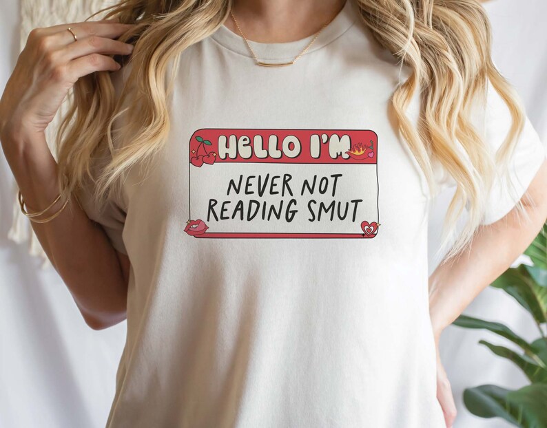May include: White t-shirt with a red name tag that says "Hello I'm Never Not Reading Smut". The name tag has cherries, a crown, and hearts on it.