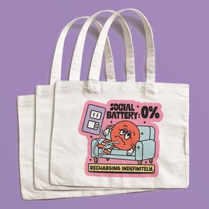May include: Three white canvas tote bags with a colorful graphic. The graphic shows a cartoon character with a red face, plugged into a wall outlet, on a blue couch. Text reads "SOCIAL BATTERY: 0% RECHARGING INDEFINITELY."