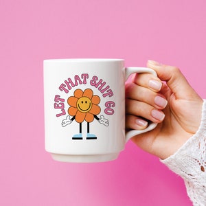 May include: White ceramic mug with a pink and orange flower graphic and the text "Let That Shit Go".