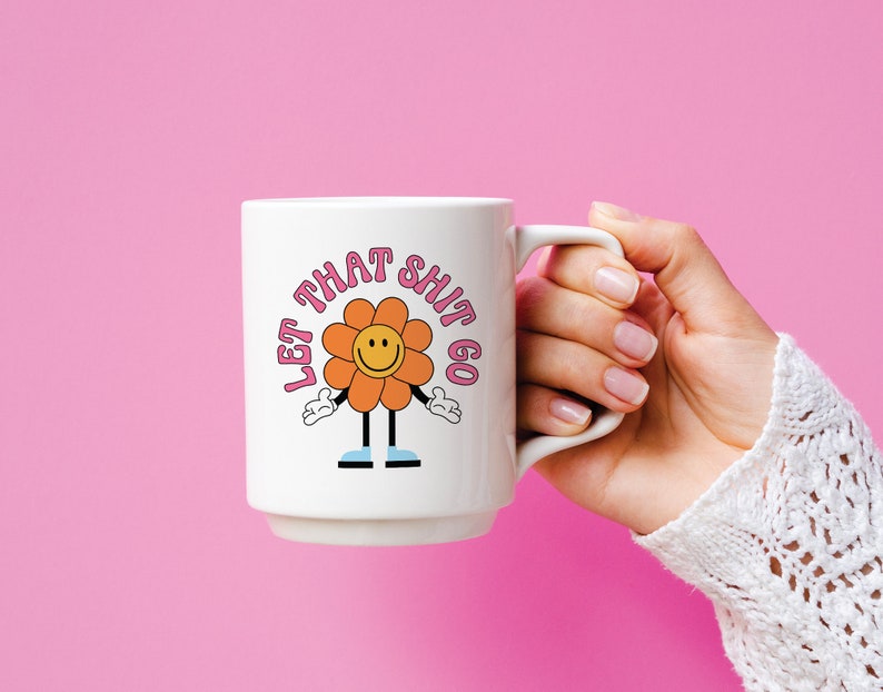 May include: White ceramic mug with a pink and orange flower graphic and the text "Let That Shit Go".