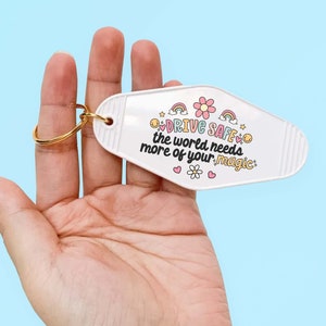 May include: White motel key chain with a gold ring. The keychain has a pink flower and rainbows with the text "Drive Safe, the world needs more of your magic."