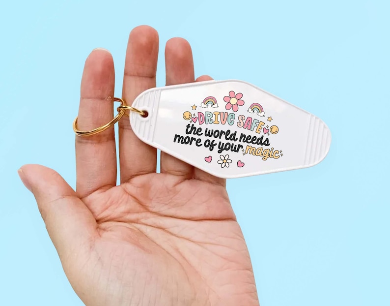 May include: White motel key chain with a gold ring. The keychain has a pink flower and rainbows with the text "Drive Safe, the world needs more of your magic."