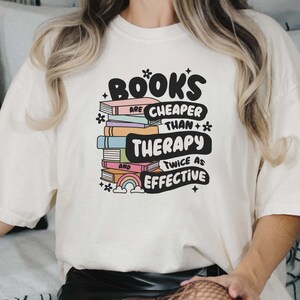 May include: White t-shirt with a graphic design of a stack of books with a rainbow and the text "Books are cheaper than therapy and twice as effective".