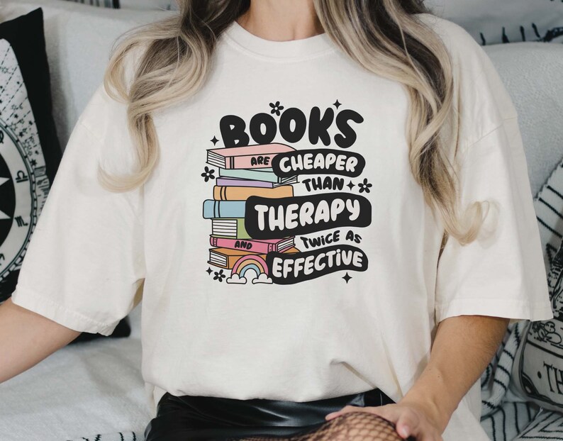 May include: White t-shirt with a graphic design of a stack of books with a rainbow and the text "Books are cheaper than therapy and twice as effective".