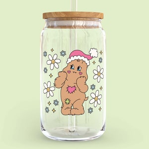 May include: A clear glass jar with a wooden lid and a straw. The jar features a cartoon bear wearing a Santa hat and surrounded by flowers.