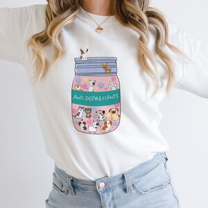 Pu&ograve; includere: Felpa bianca con una grafica di un barattolo rosa pieno di cani dei cartoni animati e il testo "Anti Depress,ants".