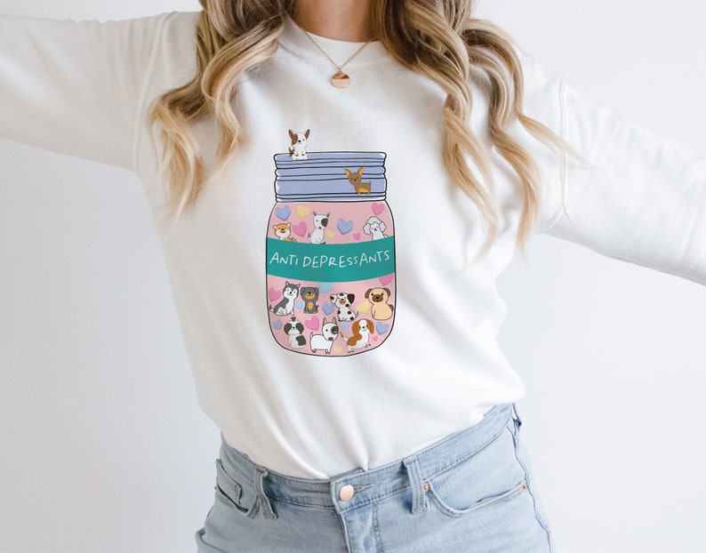 Pu&ograve; includere: Felpa bianca con una grafica di un barattolo rosa pieno di cani dei cartoni animati e il testo "Anti Depress,ants".