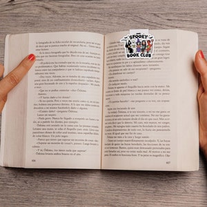 Puede incluir: Un libro abierto en las p&aacute;ginas 406-407, sostenido por manos con esmalte de u&ntilde;as rojo. Un marcap&aacute;ginas colorido "Spooky Book Club" con personajes de dibujos animados est&aacute; insertado. El libro est&aacute; sobre una superficie clara de vetas de madera.