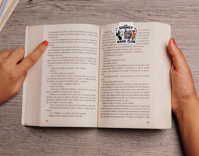Puede incluir: Un libro abierto en las p&aacute;ginas 406-407, sostenido por manos con esmalte de u&ntilde;as rojo. Un marcap&aacute;ginas colorido "Spooky Book Club" con personajes de dibujos animados est&aacute; insertado. El libro est&aacute; sobre una superficie clara de vetas de madera.
