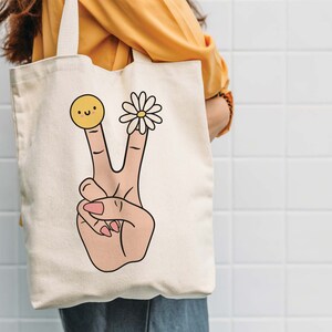 May include: A white canvas tote bag with a peace sign design. The peace sign is made up of two fingers with a smiley face on one finger and a daisy on the other.