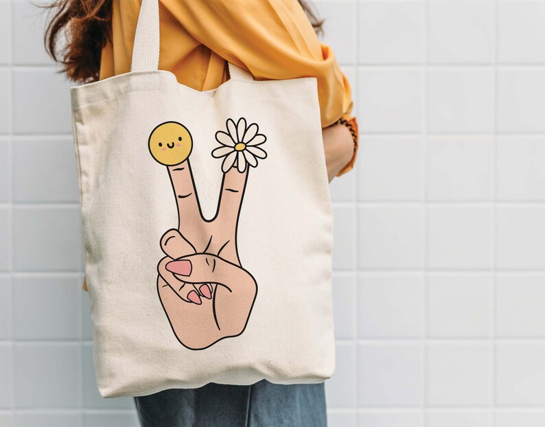 May include: A white canvas tote bag with a peace sign design. The peace sign is made up of two fingers with a smiley face on one finger and a daisy on the other.