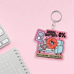 May include: A keychain with a cartoon graphic of a red character on a blue couch, plugged into a wall outlet. Text reads "SOCIAL BATTERY: 0%" and "RECHARGING INDEFINITELY." The keychain has a silver ring and is set against a pink background.