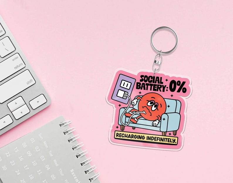 May include: A keychain with a cartoon graphic of a red character on a blue couch, plugged into a wall outlet. Text reads "SOCIAL BATTERY: 0%" and "RECHARGING INDEFINITELY." The keychain has a silver ring and is set against a pink background.