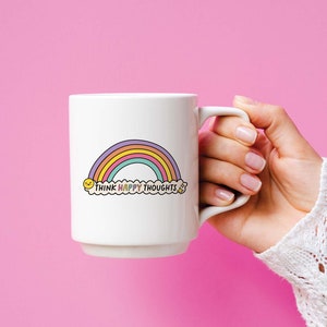 May include: White ceramic mug with a rainbow and the text "Think Happy Thoughts" in a cloud. The mug is being held by a hand wearing a white crocheted sweater.
